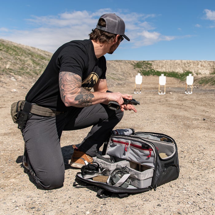 Photograph man at an outdoor shooting range inspecting a handgun near three paper targets a gray tactical bag with red accents and brown leather boots are also visible.