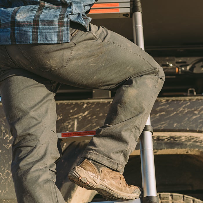 A person wearing a blue plaid shirt and mud-splattered grey pants climbs a silver telescoping ladder, a brown hiking boot visible on a rung with a red warning label