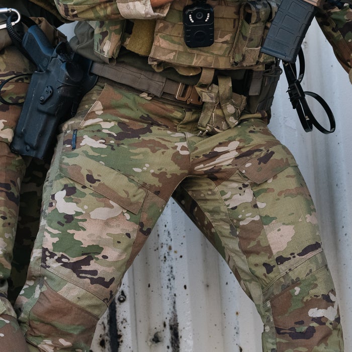 Photograph of camouflage military pants against a corrugated metal wall showing a pistol in a holster a body camera and multiple cargo pockets in shades of green brown and tan