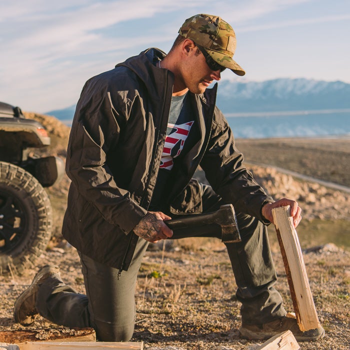 Photograph a man splitting wood with a large axe near a body of water and mountains wearing a camouflage hat and a dark jacket with an American flag design his tattoos and the golden hour light are visible