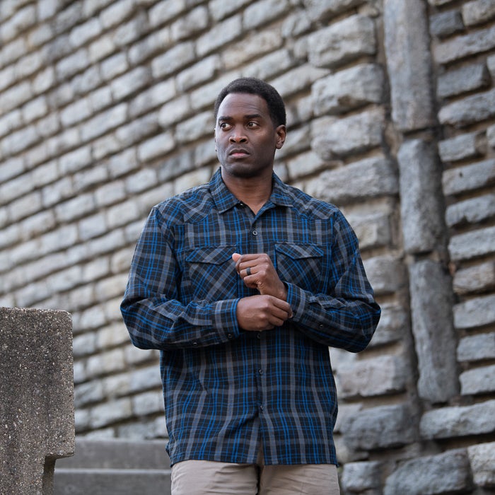 Photograph A man in a blue and gray plaid shirt stands against a stone wall adjusting his cuffs a dark ring on his finger is visible along with his serious expression and the rough texture of the bricks