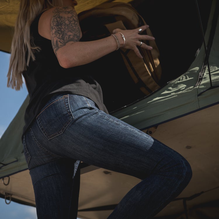 Photograph of a woman packing a tan bag into a rooftop tent showcasing dark wash jeans floral arm tattoo and silver bracelets against a bright blue sky