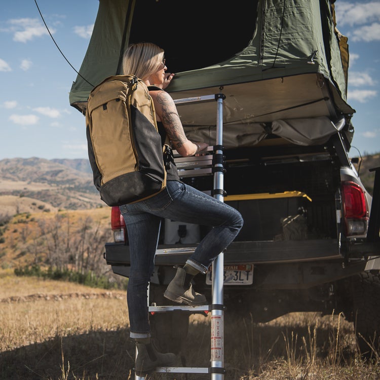 Photograph Woman climbing ladder to rooftop tent on truck in mountainous landscape featuring tan backpack brown boots and blue jeans
