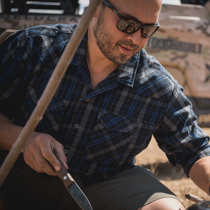 Photograph of a man wearing a blue and gray plaid shirt sharpening a knife outdoors near a vehicle with a blue and gray plaid shirt, a wooden stick, and reflective sunglasses