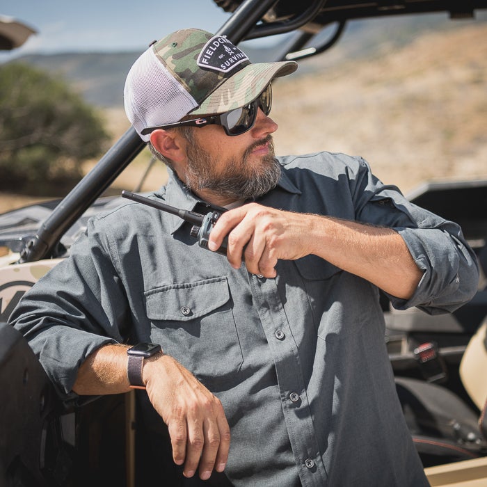Photograph of a man using a two-way radio near an off-road vehicle in a desert landscape featuring a camouflage hat, a smartwatch, and a sunny sky