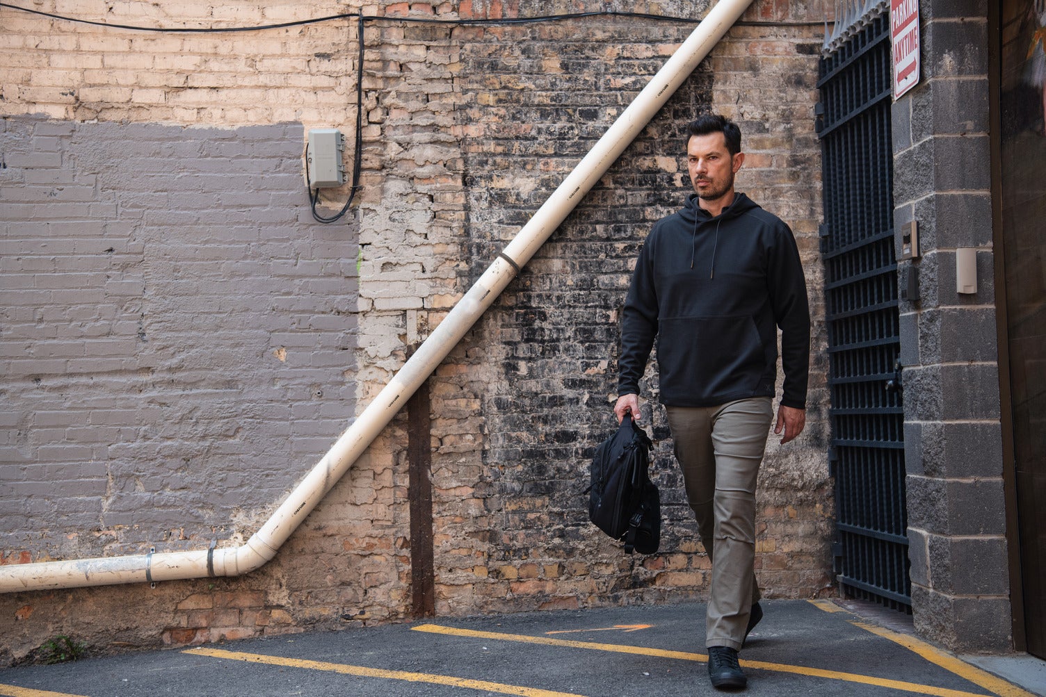 Photography a man walking down an alleyway with exposed brick walls a large white pipe and a black metal gate featuring grey painted brickwork and a black backpack