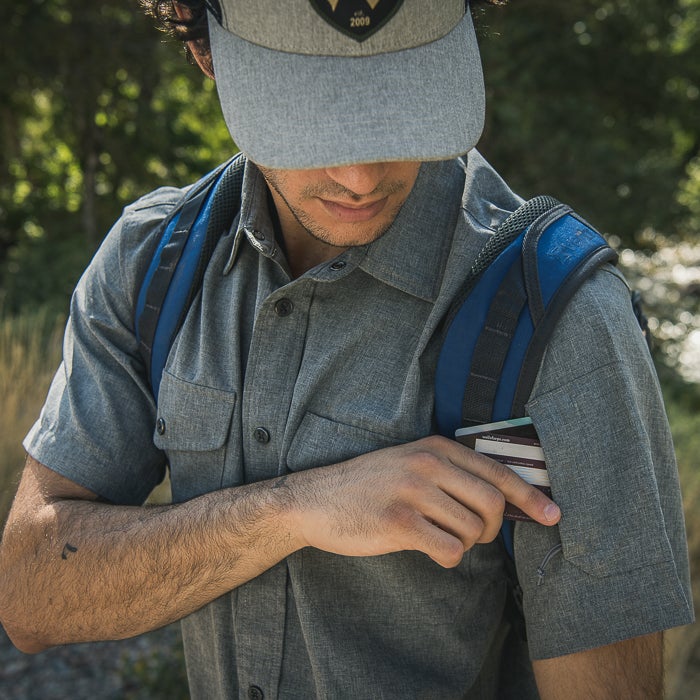 Photography a man in a gray shirt and hat accessing a pocket outdoors near green trees and a flowing stream featuring a blue backpack a small tattoo and a credit card.