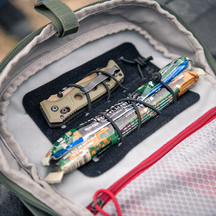 Photograph of a tan and black folding knife and two camouflaged energy bars inside a grey and green tactical bag featuring red accents and black securing cords