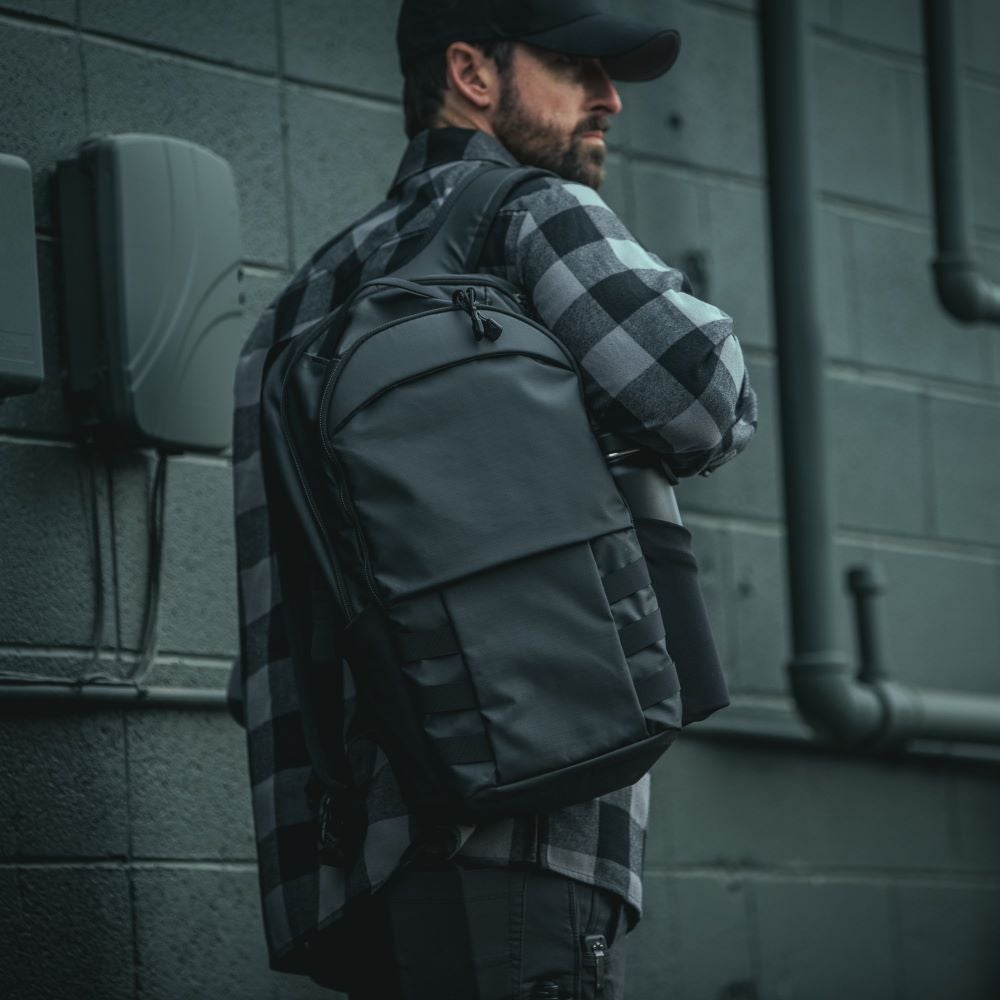 Photography of a man wearing a gray and black plaid shirt and carrying a sleek black backpack against a gray urban wall featuring visible pipes and electrical boxes