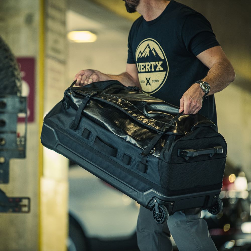Photograph of a man in a parking garage carrying a large black VERTX wheeled duffel bag with rugged handles and textured fabric featuring a mountain logo on his shirt