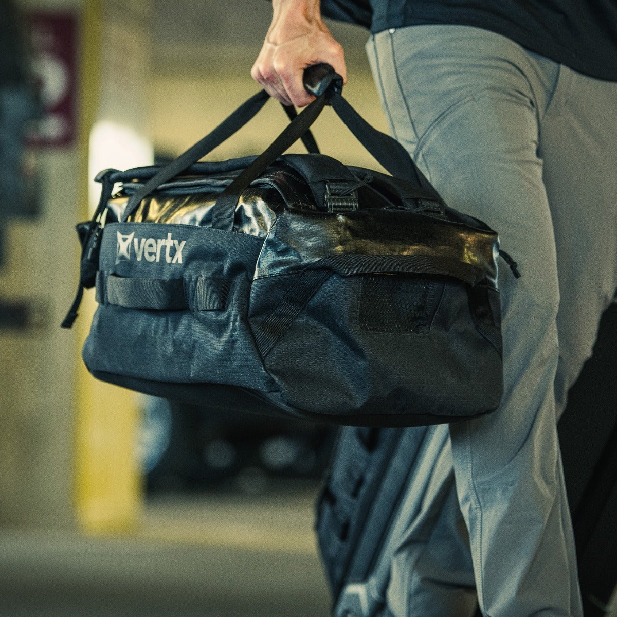 Product Photography: A person carries a black Vertx duffel bag with gray accents and durable straps through a dimly lit parking garage featuring a yellow pillar and part of a wheeled suitcase