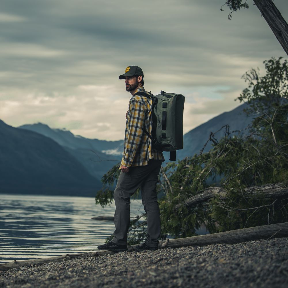 Photograph of a man with a green backpack standing by a lake in a mountainous region with a yellow and black plaid shirt dark gray pants and a dark green hat near rocky shoreline and imposing mountain range