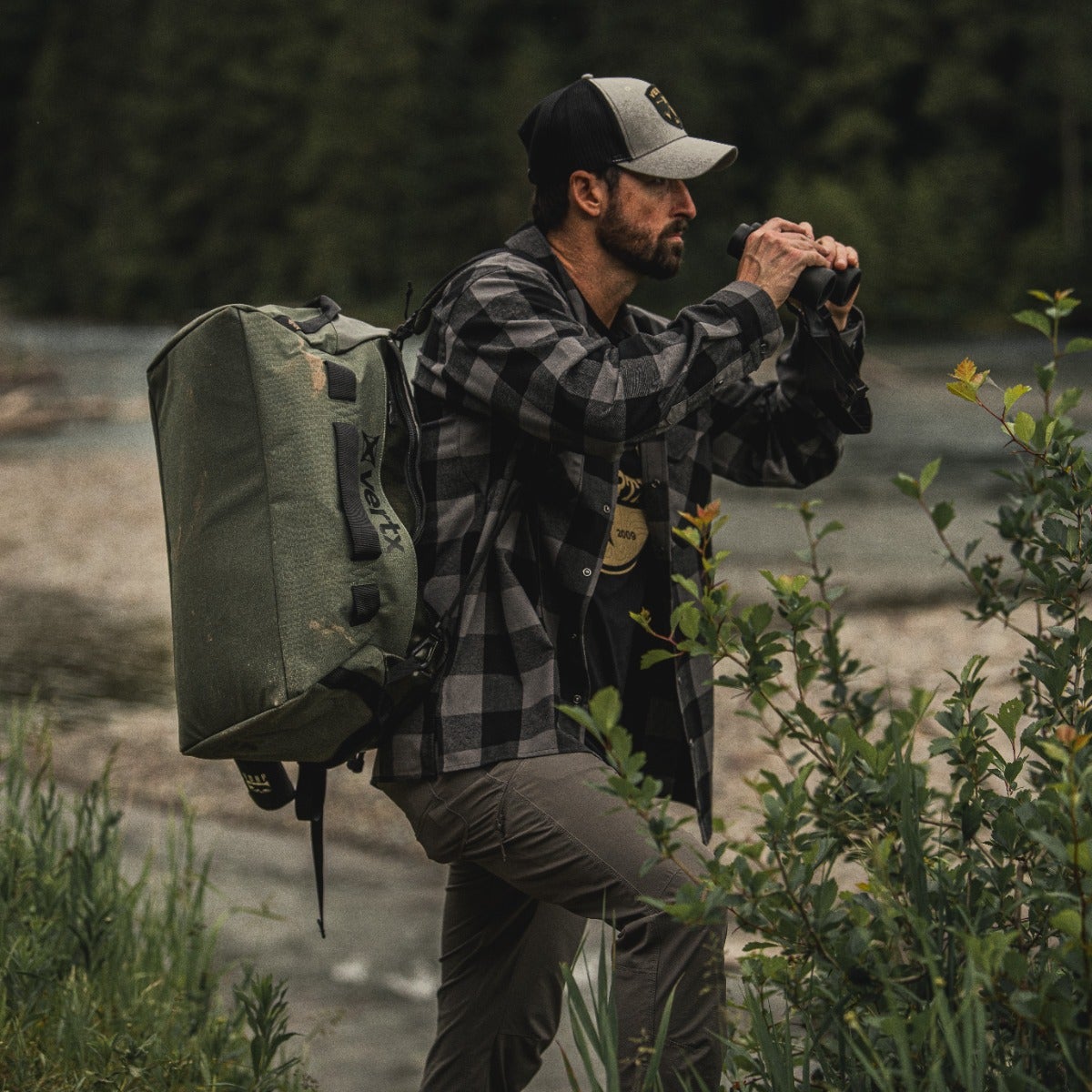 Photography Man with Vertex backpack using binoculars by a river with lush green vegetation and a gray-green, olive-toned bag a black and gray flannel shirt