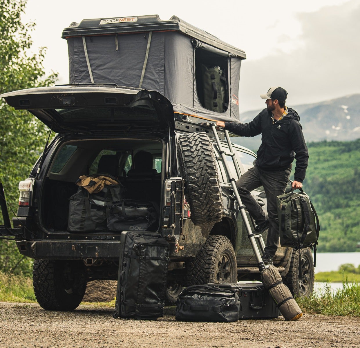 Contemporary photograph of a man near a lake and mountains with a black Toyota Land Cruiser, a Roofnest rooftop tent, and numerous black duffel bags, showcasing rugged tires, a dark gray tent, and olive green backpack.