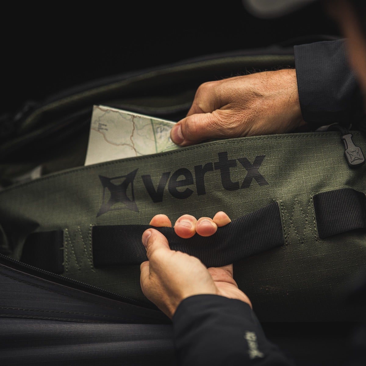 Product shot showing hands interacting with a Vertx olive-green duffel bag featuring a black logo and sturdy black straps a map is partially visible inside