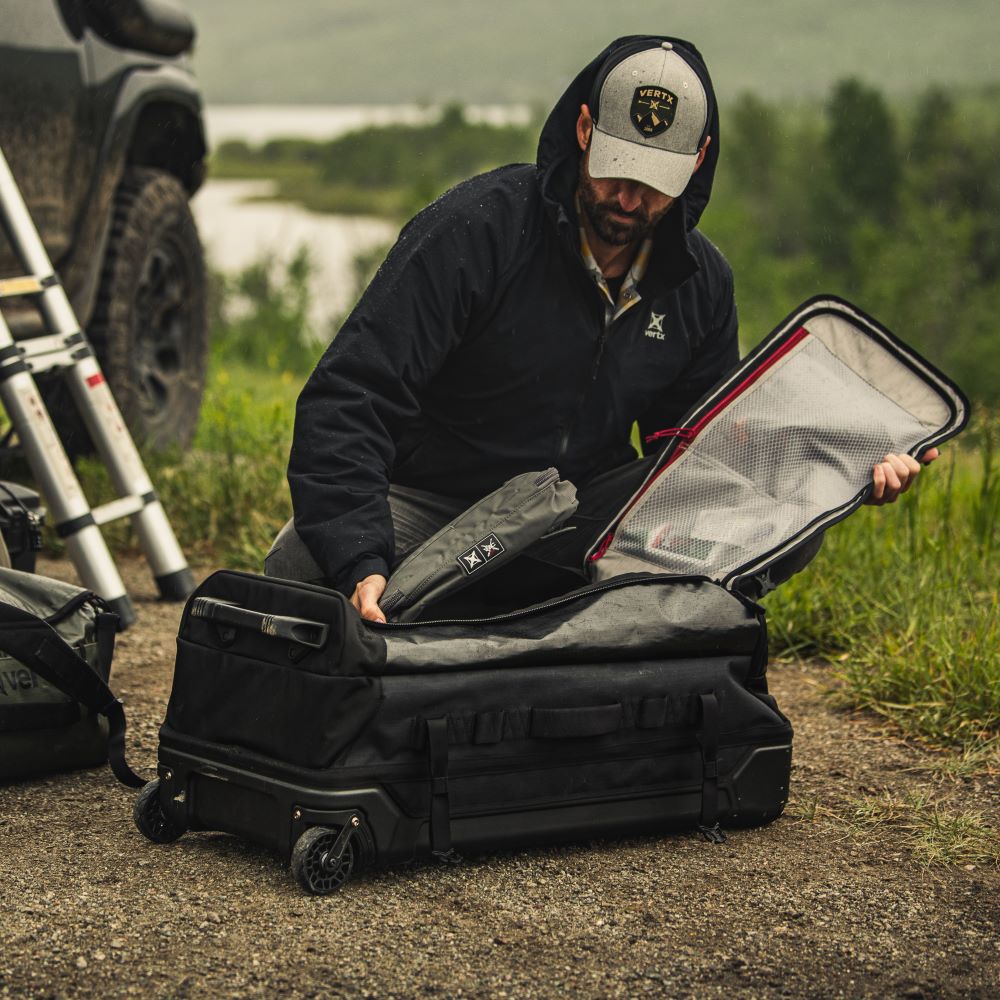 Photograph of a man packing a large black wheeled duffel bag outdoors near a body of water and a grey SUV featuring red accents and a grey Vertix hat.