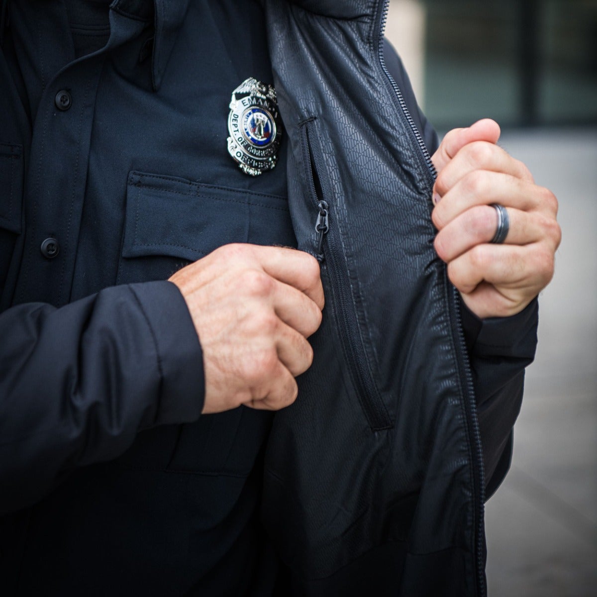 Photograph showing a close-up of a person wearing a dark jacket and shirt with a silver EMA Department of Emergency Management badge featuring an eagle and American flag detail, a visible zipper, and a textured fabric with subtle diamond pattern.