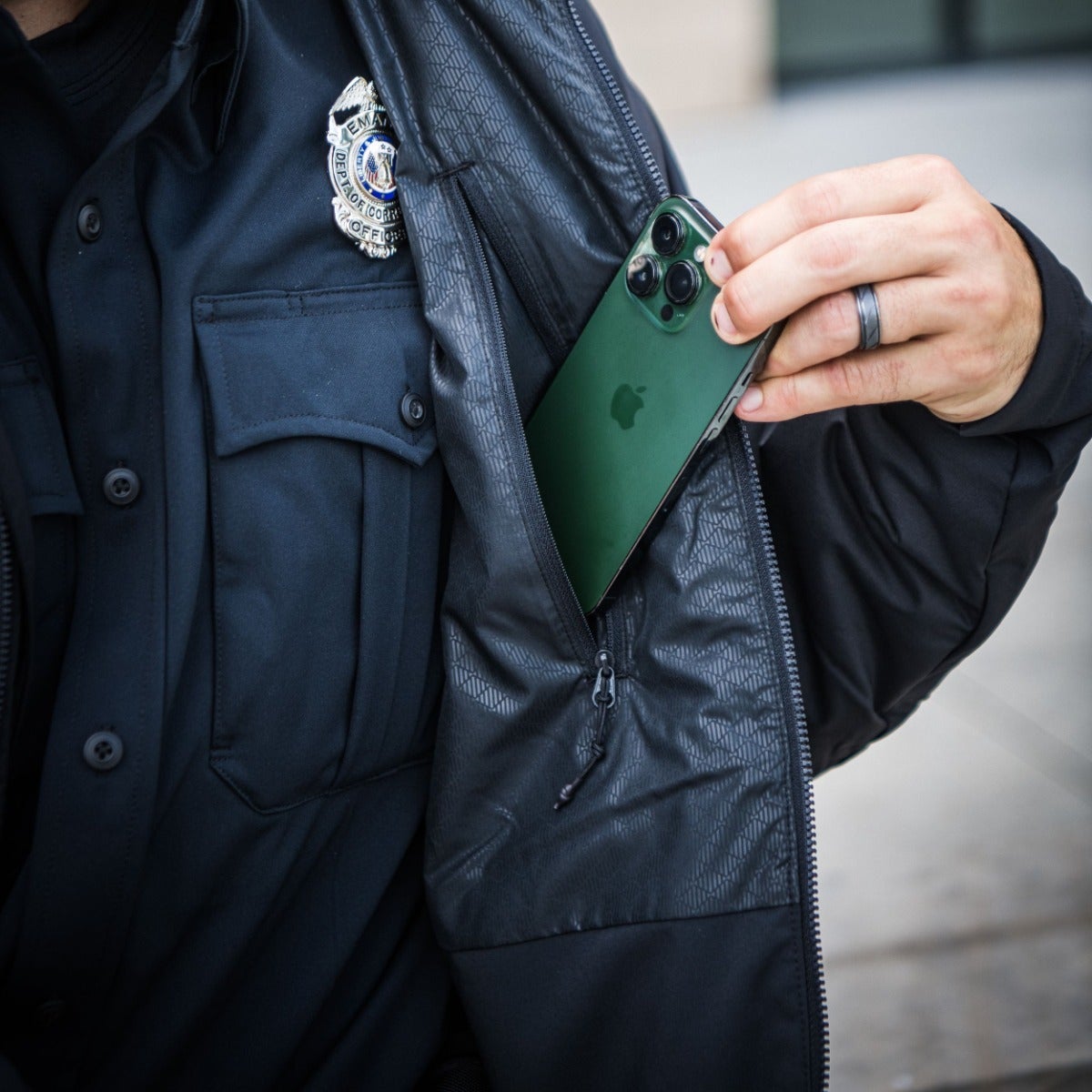 Photograph showing a law enforcement officer placing a dark green Apple iPhone 12 Pro Max in a jacket pocket the officer's badge is visible along with a textured fabric and a silver ring