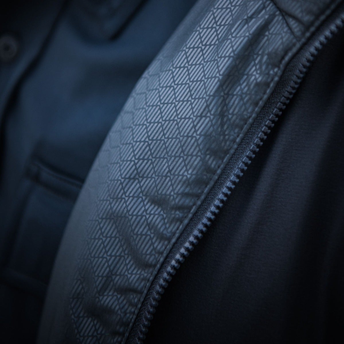 Close-up photograph detail of a dark blue jacket featuring a lighter gray geometric patterned panel and a visible zipper