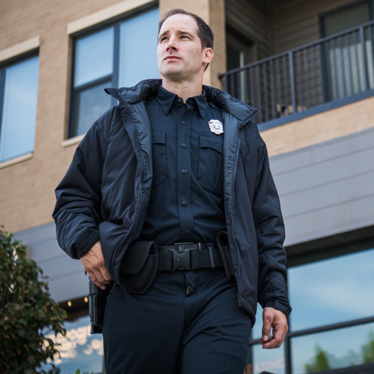 Photograph of a police officer in a dark uniform and jacket outside a modern building with large windows and a balcony, a silver badge, and a black utility belt.
