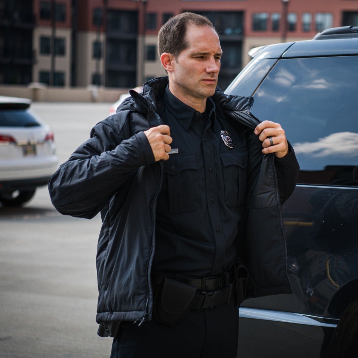 Photograph of a police officer putting on his jacket in a city parking lot near an apartment building and a dark SUV featuring a black uniform a shiny badge and a tactical belt