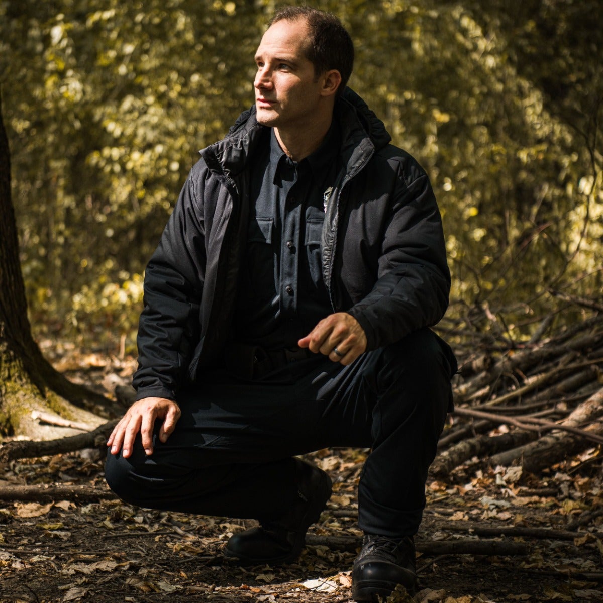 Photograph of a man in a black jacket and pants, crouching in a sun-dappled forest with a large tree trunk and fallen branches exhibiting warm autumnal colors and dappled light
