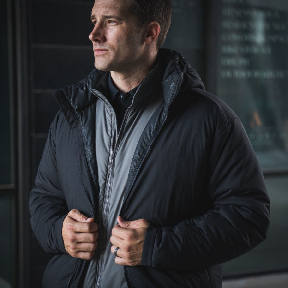 Photography unknown artist man in black puffer jacket with gray inner layer and dark ring standing outdoors near modern glass building with blurred background details