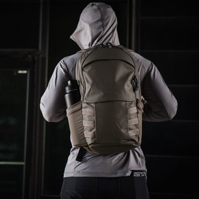 Photography Unknown Artist a person wearing a gray hooded sweatshirt and a muted olive-green backpack with side compression straps and a black water bottle in a side pocket stands against a dark background