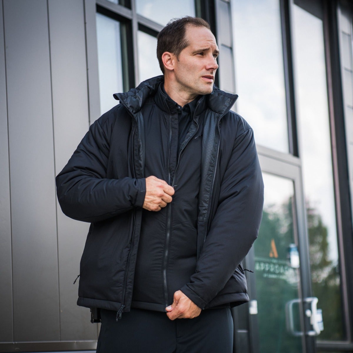 Photograph of a man in a black insulated jacket adjusting the zipper outside a modern building with large glass windows and gray metal siding