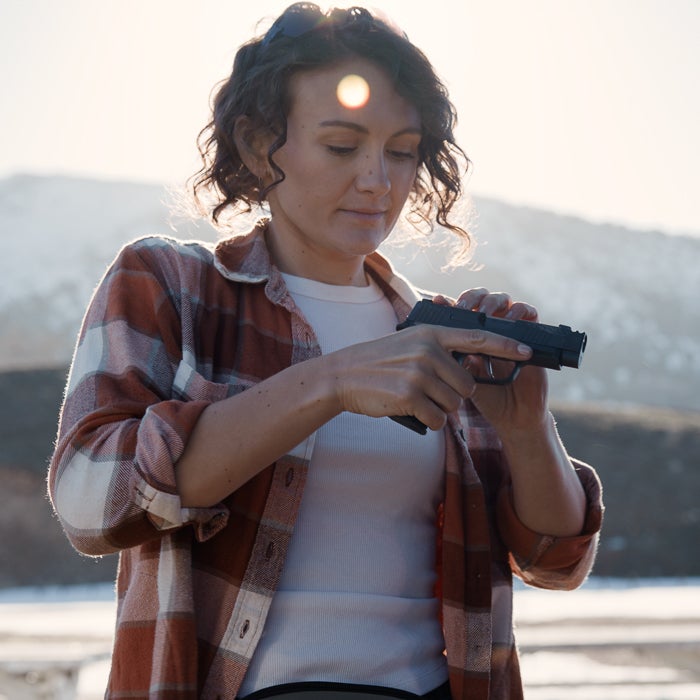 Photograph of a woman in a red and white plaid shirt examining a handgun against a mountainous backdrop with sun flare and lens highlights