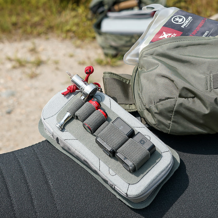 Product photography showing a gray zippered pouch containing a silver tool, red and gray battery packs, and other accessories attached to a camouflage backpack outdoors