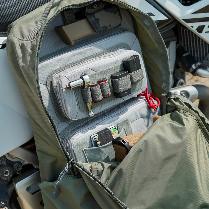 Photograph of an olive-green backpack's organized interior showing various tools, a firearm, and gray pouches next to a motorcycle