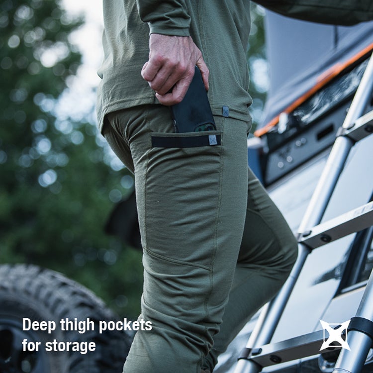 Photograph showing a person wearing olive-green merino wool pants with deep thigh pockets and a phone in one pocket near a vehicle and an extending ladder featuring dark gray fabric, black pocket trim, and a blurred green background