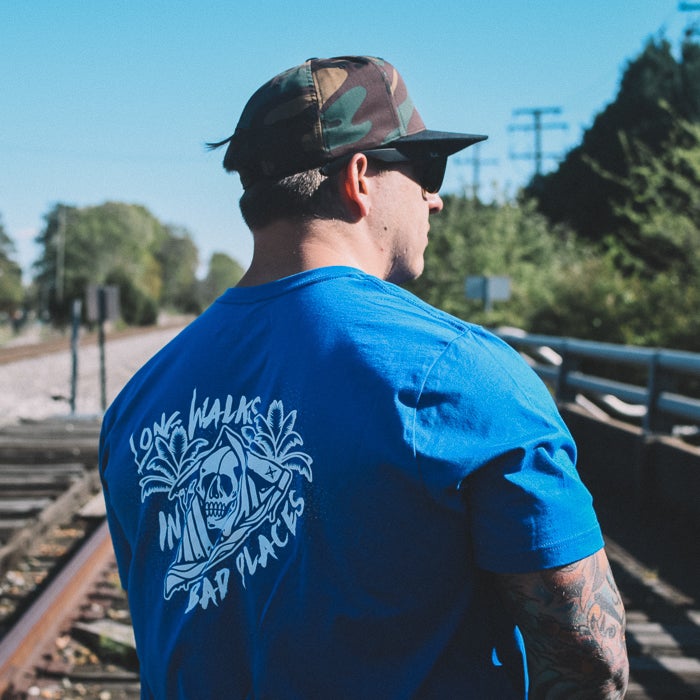Photograph of a man wearing a royal blue t-shirt featuring a skull graphic and camouflage hat standing on train tracks near lush green trees and power lines showcasing a vibrant design with palm trees and a unique skull motif