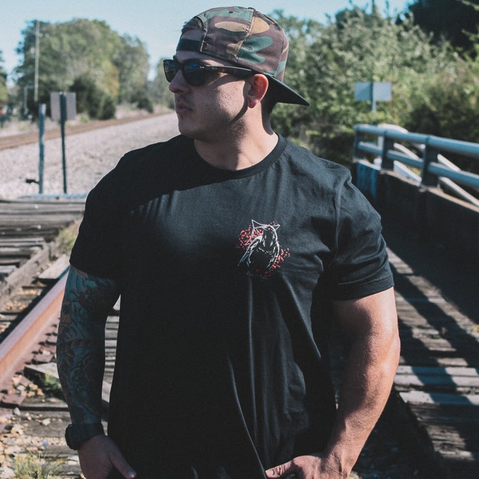 Photography model wearing a black t-shirt featuring a stylized crow with red accents near railroad tracks and a bridge showcasing a faded camo hat and tattooed arms