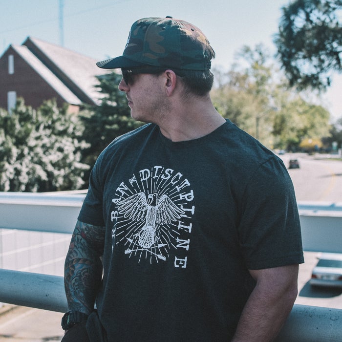 Photography Unknown Artist A man wearing a dark gray t-shirt featuring a stylized eagle graphic and the words BRAVE DISCIPLINE stands on a bridge with a brick building and lush green trees in the background the eagle is encircled by sun rays and arrows and the man sports a camouflage hat and tattoos