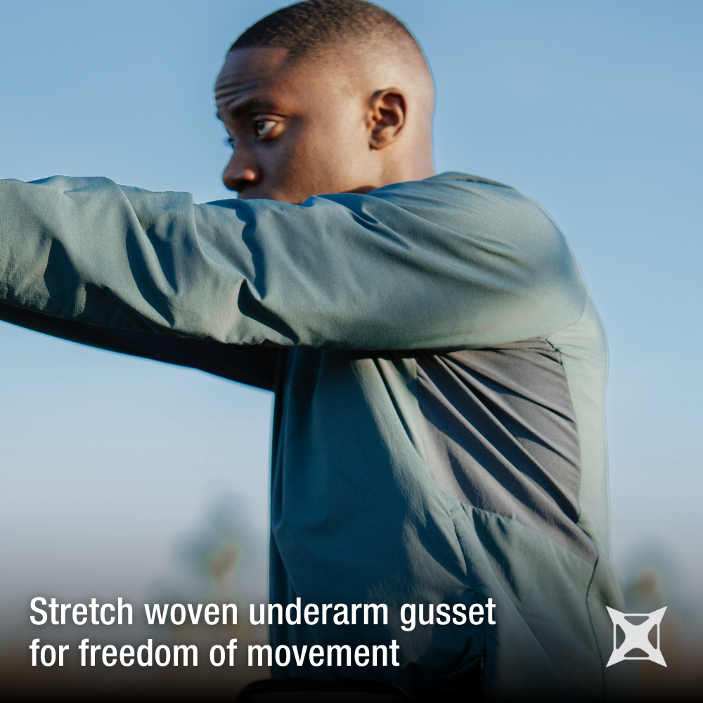 Photograph of a man wearing a teal athletic jacket with gray underarm panels outdoors against a bright blue sky showing off the garment's stretch woven fabric and freedom of movement