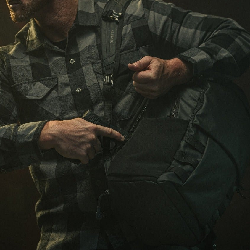 Photograph man in gray and black plaid shirt accessing handgun in black backpack with Xferty logo and compression straps against dark background