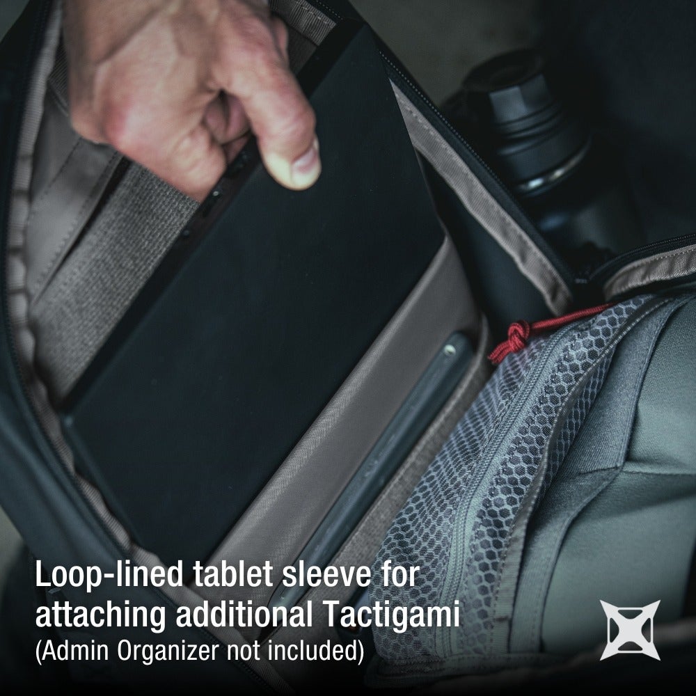 Product photography showing a hand inserting a black tablet into a beige loop-lined sleeve inside a gray and black backpack with a red zipper pull and gray honeycomb patterned pocket