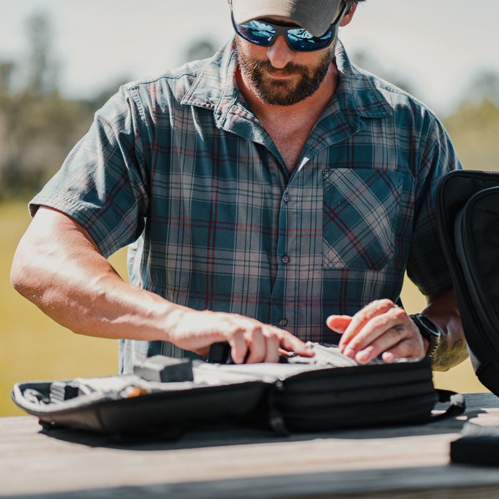 Photograph of a man outdoors near grassy fields, meticulously organizing gear in a black case; he wears a blue and red plaid shirt, sunglasses, and a khaki cap