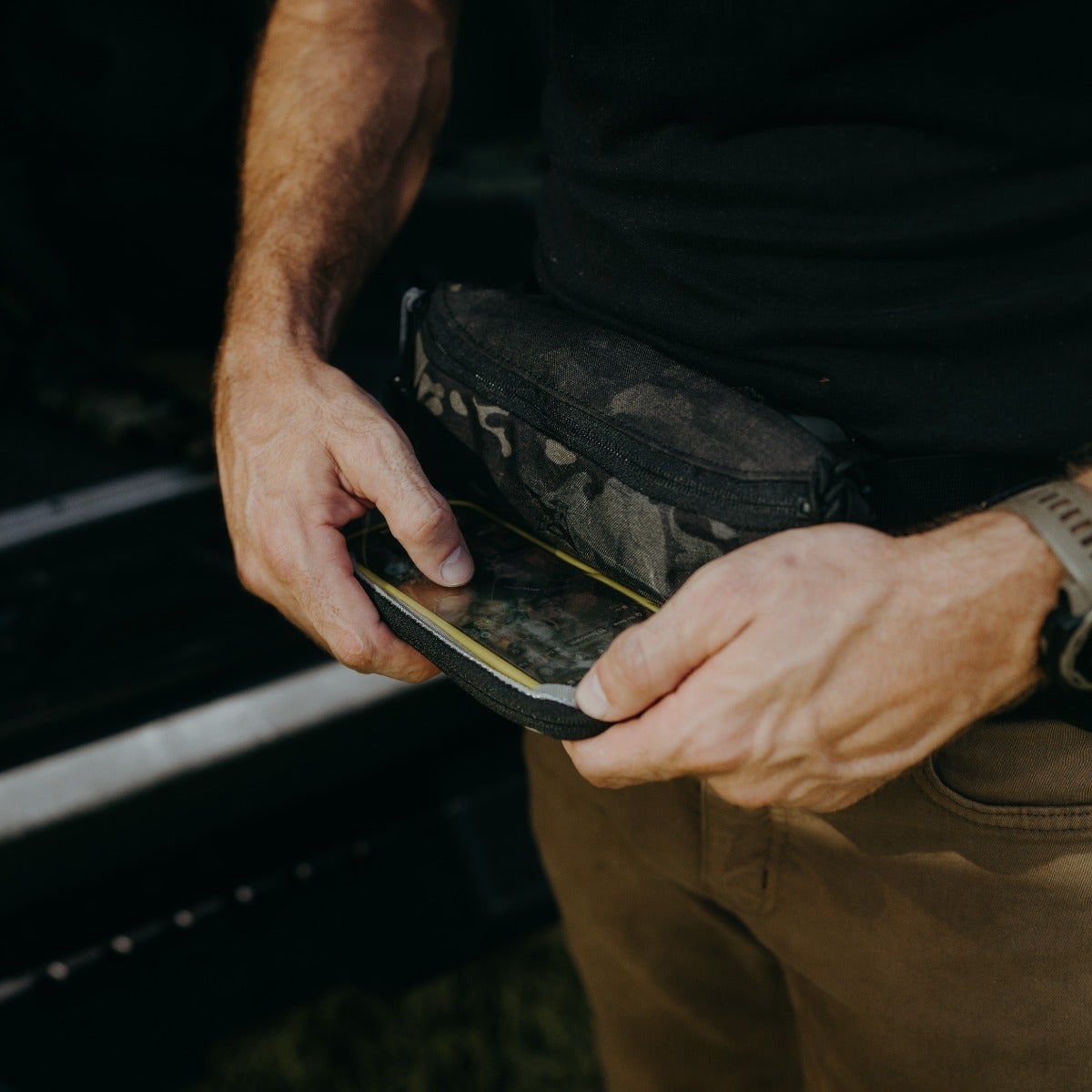Photograph of a person's hands removing a smartphone from a black and camouflage fanny pack with a yellow waterproof case detail khaki pants and a watch are visible