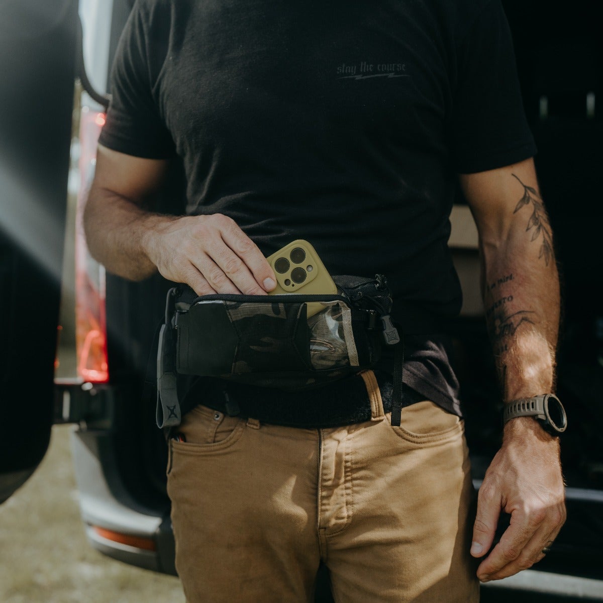 A photograph showing a man placing a olive-green iPhone into a camouflage fanny pack worn around his khaki pants near a vehicle with a visible red taillight and the man's arm showing a leaf tattoo and a dark-colored smartwatch