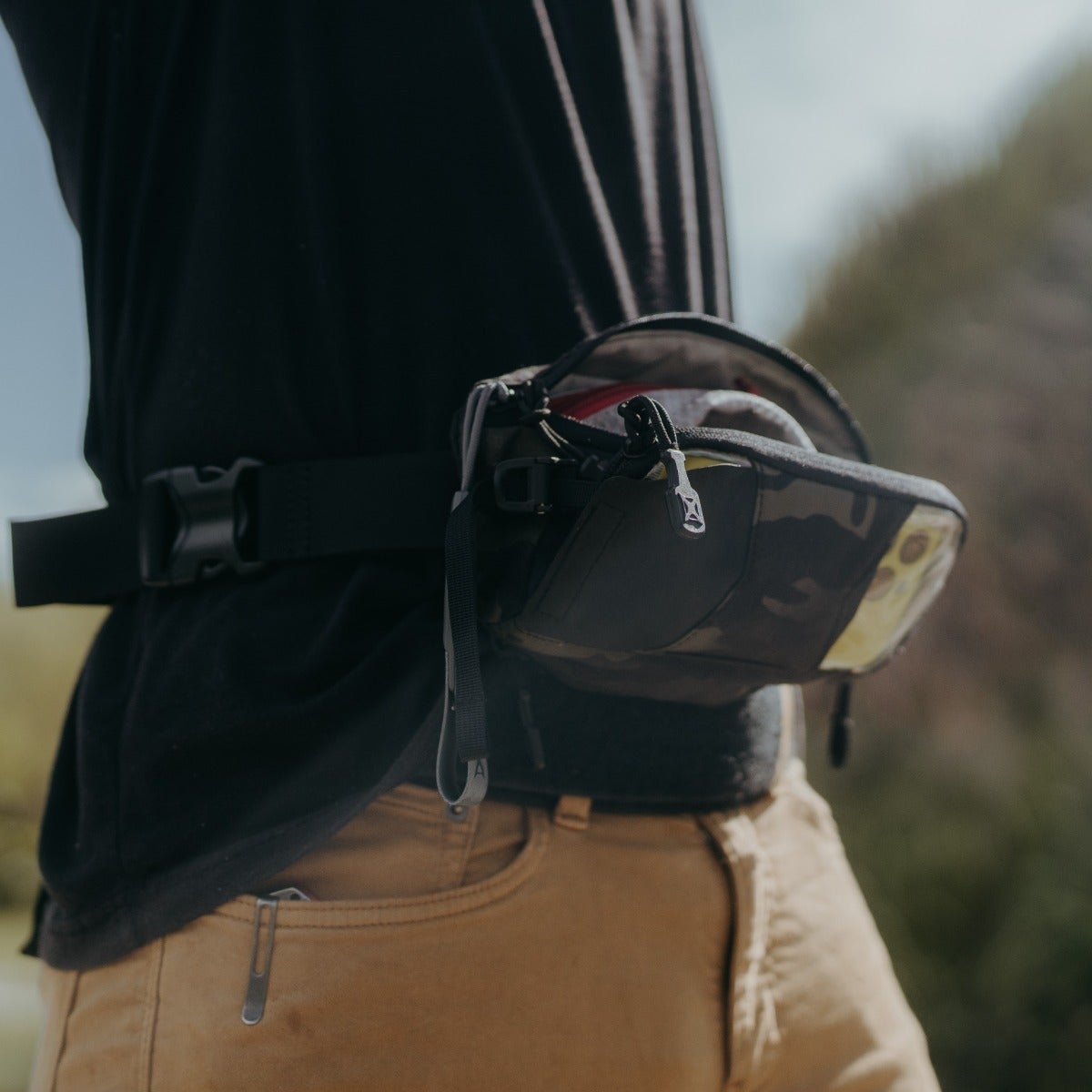 Photograph of a person wearing a black and gray fanny pack with a unique zipper pull and yellow accents against a blurred outdoor background featuring muted greens and browns.
