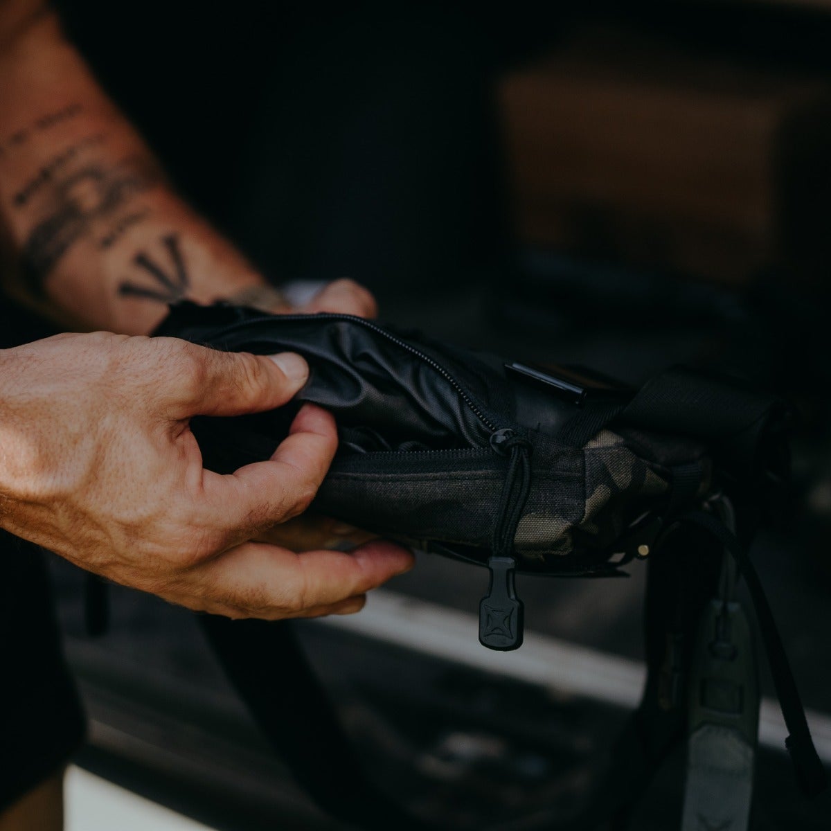 Photograph a person's hands interacting with a black and camouflage patterned bag featuring a unique zipper pull and inner waterproof liner near a dark-colored vehicle