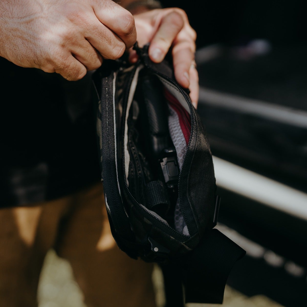 Photograph showing a person unpacking a camouflage fanny pack containing a handgun a red zipper pull and gray mesh interior pocket