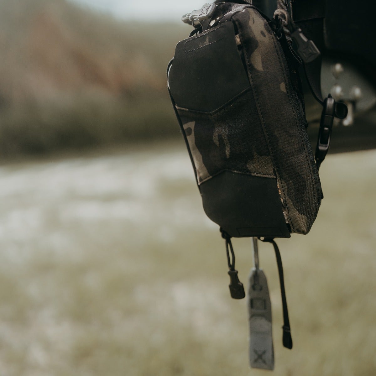 Photography of a camouflage and black utility pouch with grey accents hanging outdoors against a blurred background of muted greens and browns.