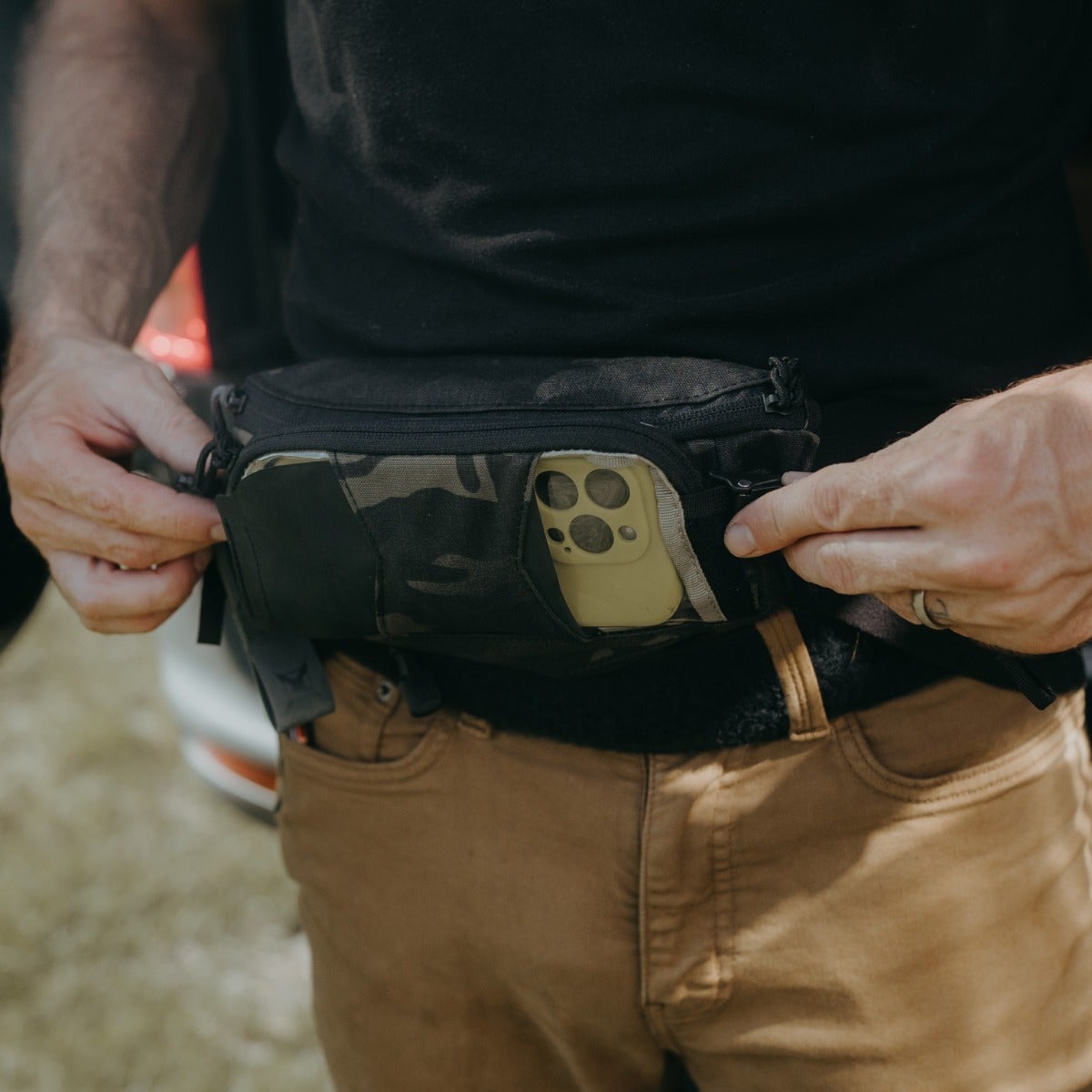 Photograph of a person wearing a black and camouflage fanny pack with an olive green iPhone visible in a dedicated pocket the pack is fastened around khaki colored pants near a blurred car background