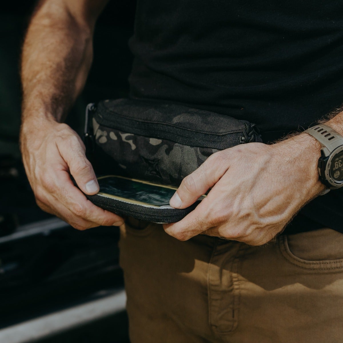 Photograph showing a person's hands holding a black and gray camouflage fanny pack containing a smartphone with a beige lining near a vehicle with khaki pants and a Garmin watch