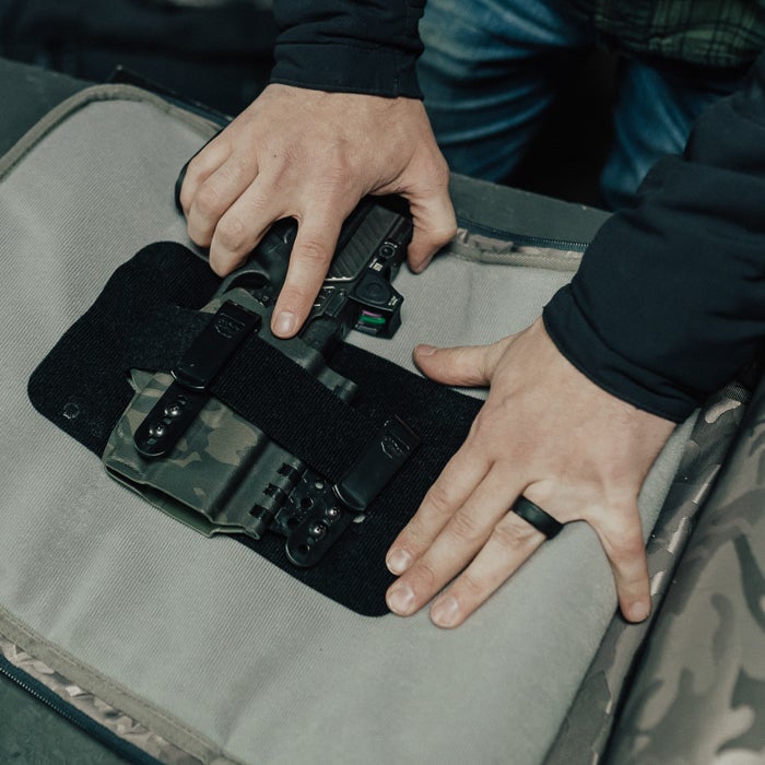 Photograph showing a person's hands placing a handgun with a red dot sight into a camouflage-patterned holster inside a gray padded case featuring black straps and metal accents
