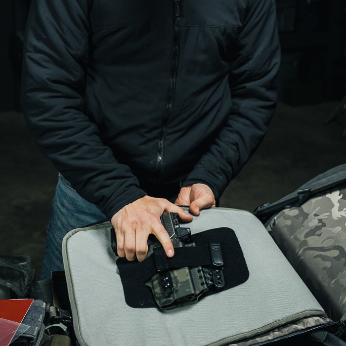 A photograph shows a person placing a handgun with a camouflage holster into a grey padded bag inside a dark room.
